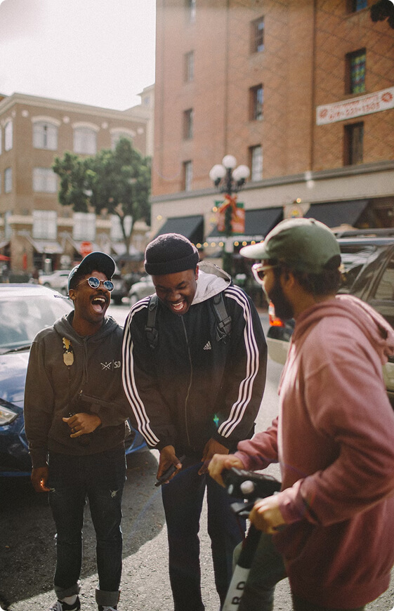Three people laughing on the street, one in a black hoodie, another in striped jacket, third in pink sweater.