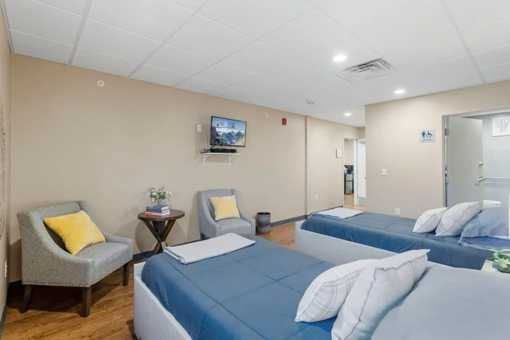 Room with two beds, gray chairs, yellow pillows, TV on wall, wooden floor, and beige walls.