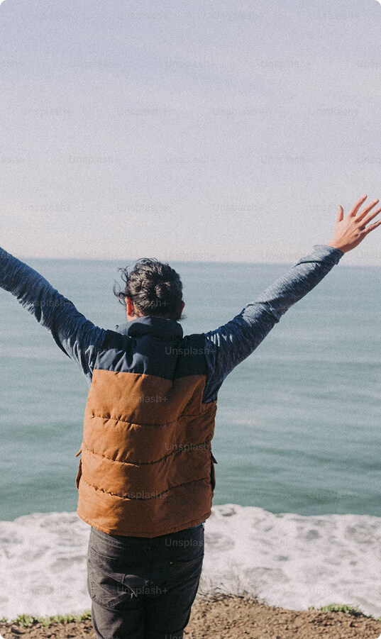 Person in a brown vest standing with arms outstretched facing the ocean.