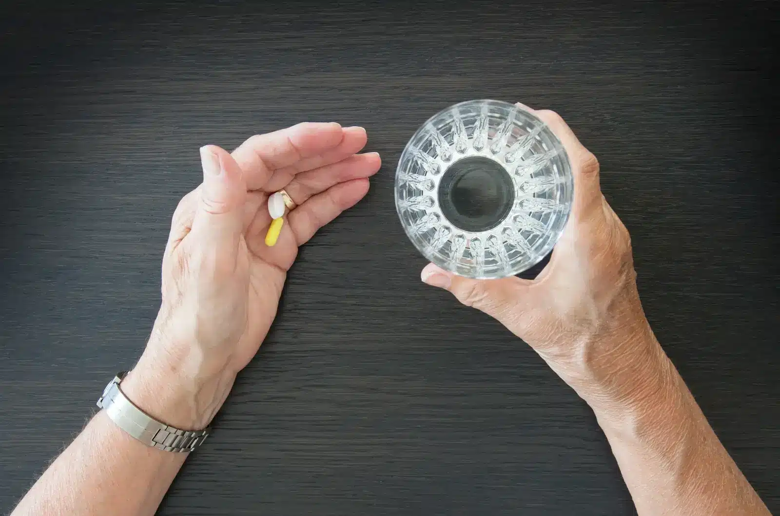 Person holding pill in left hand and glass of water in right hand.