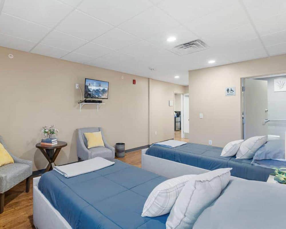 Two beds with blue covers, chairs with yellow pillows, and a wall-mounted TV in a beige room.