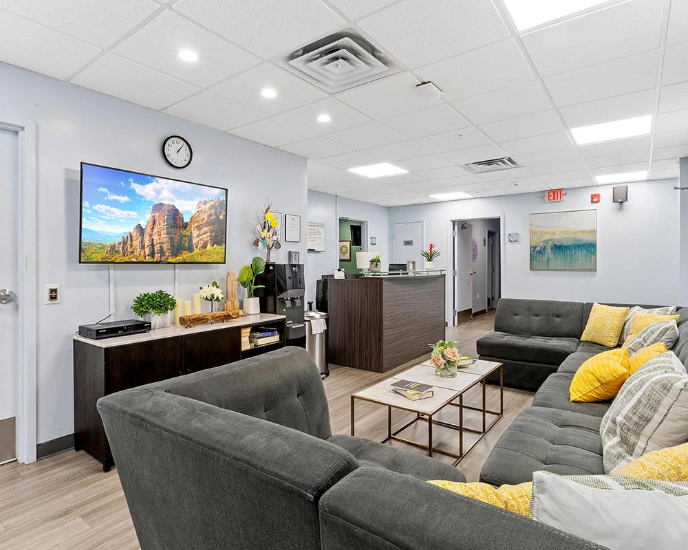 Reception area with gray couches, yellow pillows, wall art, TV, and plants.