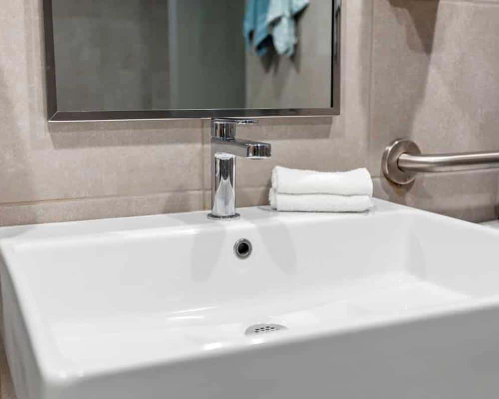 Bathroom sink with chrome faucet, folded white towels, and a safety grab bar on tiled wall.