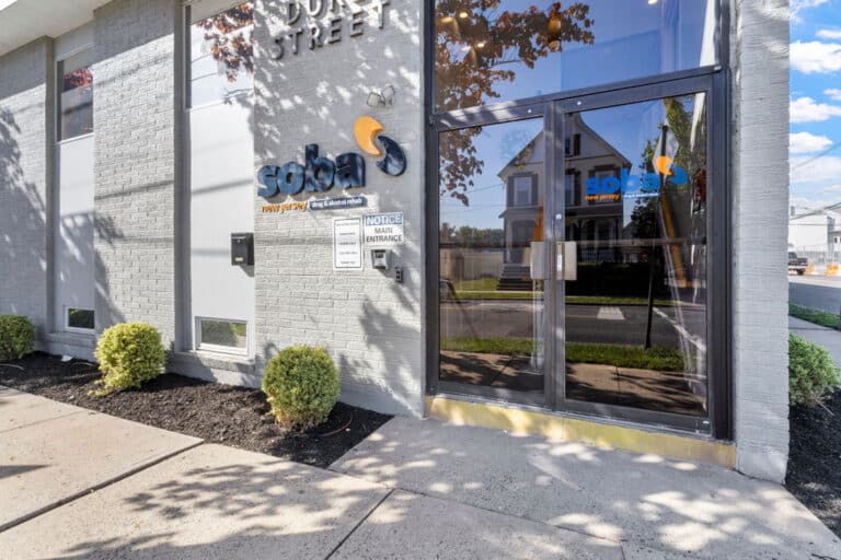 Building entrance with soba new jersey logo on gray wall, glass door reflecting street view, and small bushes.
