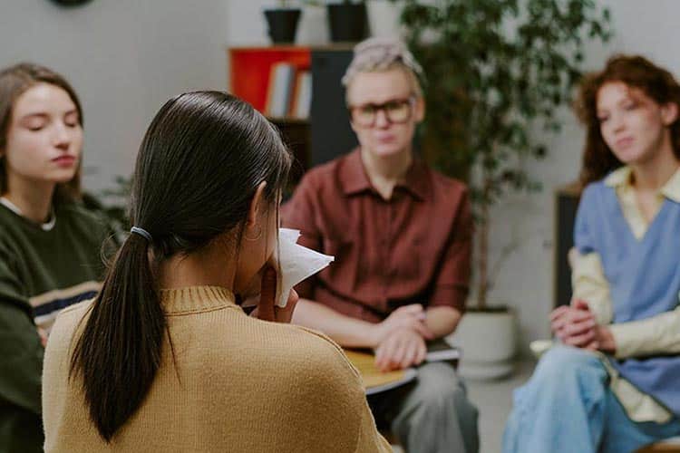Person in brown sweater holding tissue in group therapy session.