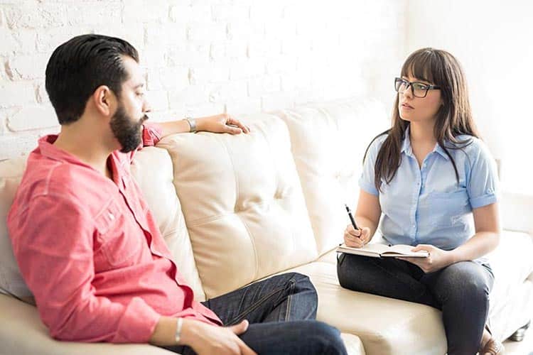 Person in a light blue shirt taking notes, speaking with a man in a red shirt on a couch.