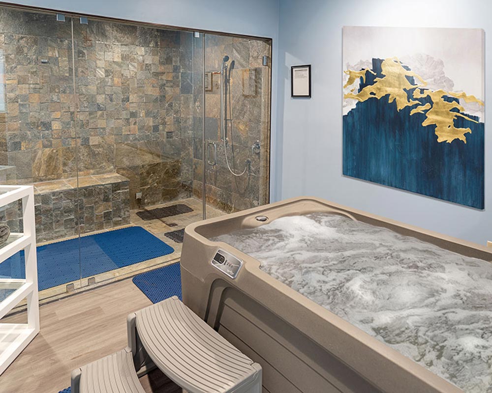 Spa room with hot tub, glass shower enclosure, abstract painting, and blue mats.