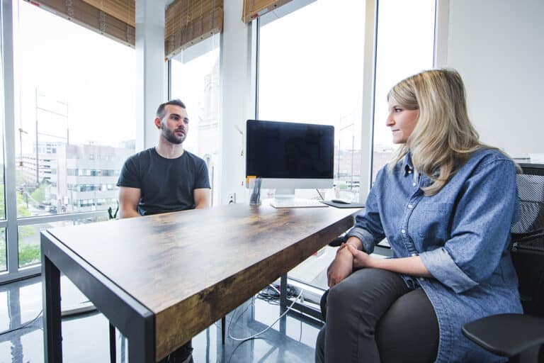Person in a black shirt talking to woman in denim shirt at a wooden table.