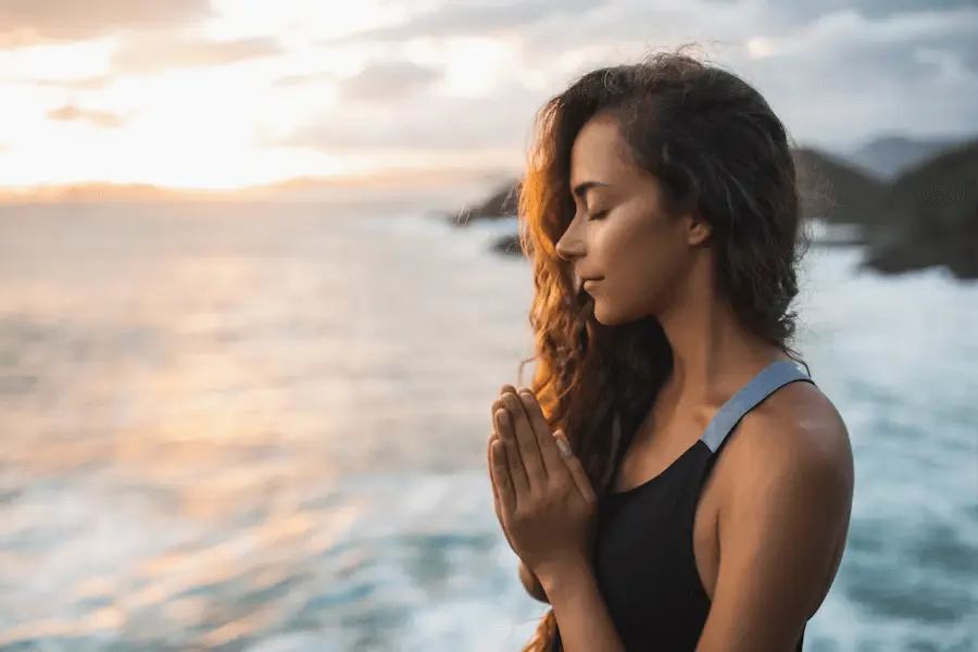 Person with long hair in black tank top meditating by the ocean at sunset.