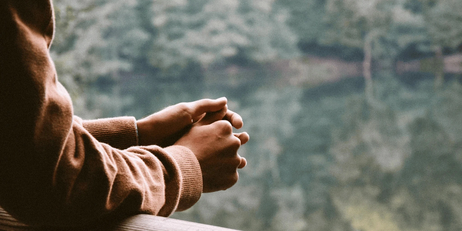 Person wearing a brown sweater standing by a railing with hands clasped.