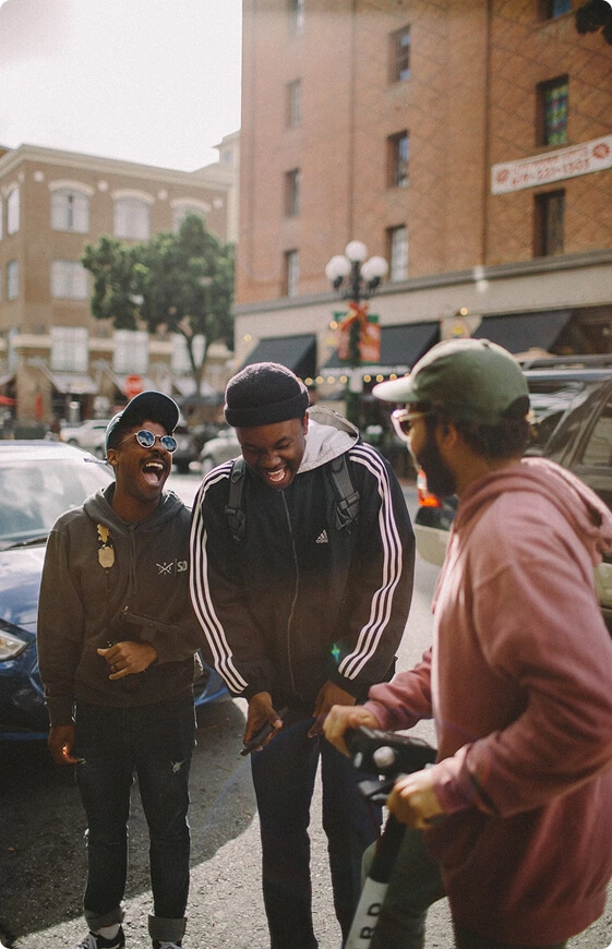Three people laughing on the street, one in a black hoodie, another in striped jacket, third in pink sweater.