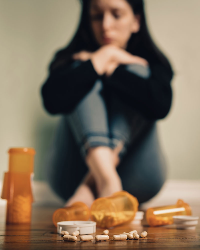 Person sitting with crossed legs, blurred face, near scattered pill bottles on the floor.