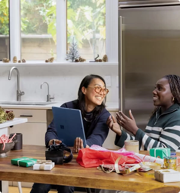 Two people at a kitchen table, one with glasses holding a blue laptop, both smiling and talking.